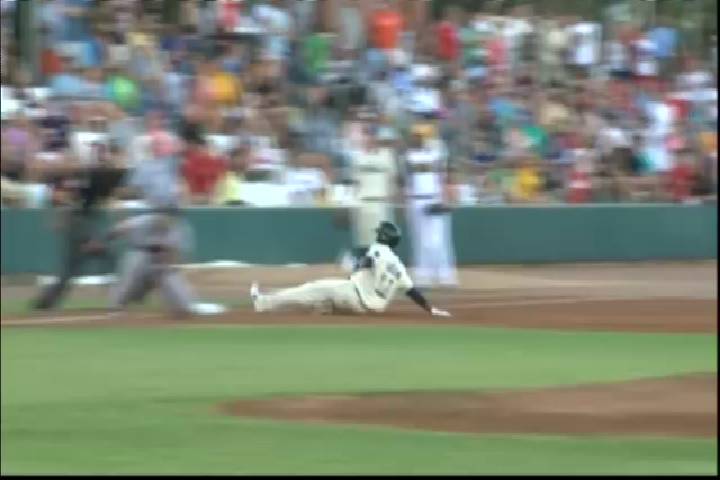 Blurred action shot of a baseball player sliding across infield dirt, with a crowd in colorful clothing blurred in the background, energetic and warm in tone